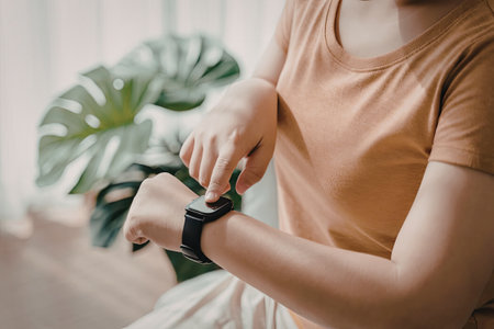 A person wearing a brown t-shirt is adjusting a smartwatch on their wrist. The scene is set indoors with a large green leafy plant in the background.の写真素材