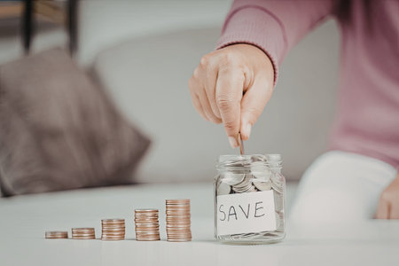 A person is shown adding coins to a glass jar labeled 'SAVE'. The jar is surrounded by stacks of coins on a table, indicating a theme of saving money.の写真素材