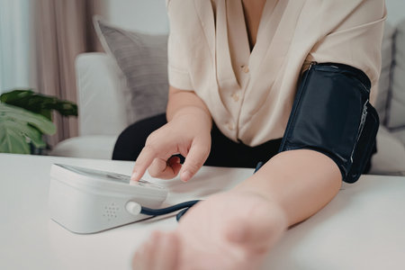 A person is seated on a couch, using a digital blood pressure monitor to check their blood pressure...