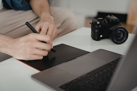 A person is seated at a table, drawing on a digital tablet with a stylus. A laptop and a camera are placed nearby on the table.の写真素材