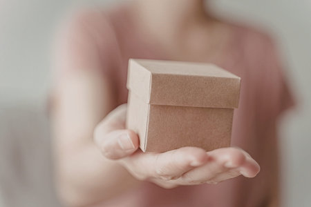 A close-up image of a person holding a small brown box in their hands. The person is wearing a pink...