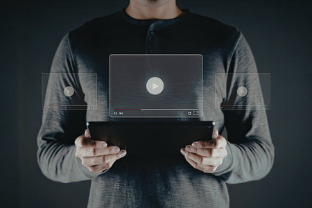 A person is holding a tablet device in their hands, displaying a video playing on the screen. The individual is wearing a dark-colored sweater and is positioned against a dark background.の写真素材