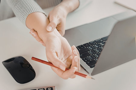 An image showing a person holding their hand in pain while sitting near a laptop and computer mouse.