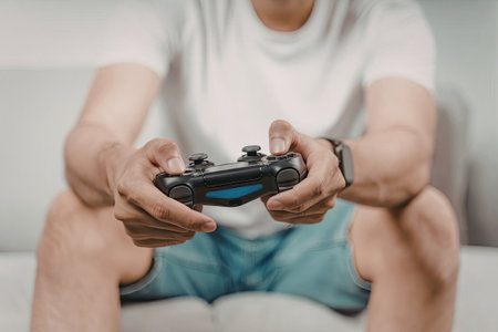 A person is sitting on a couch, intently playing a video game using a black game controller. The individual is wearing a white t-shirt and blue shorts.の写真素材