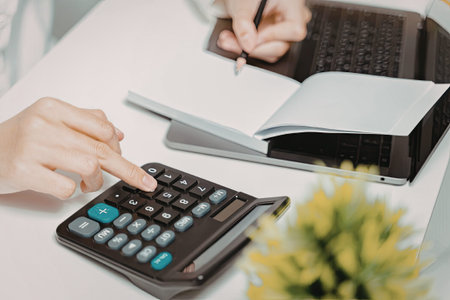 A close-up image of a person's hand using a calculator on a desk. The desk also has a laptop, a...