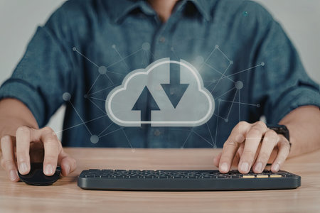 A person is seated at a desk using a computer with a cloud download icon visible on the screen. The...
