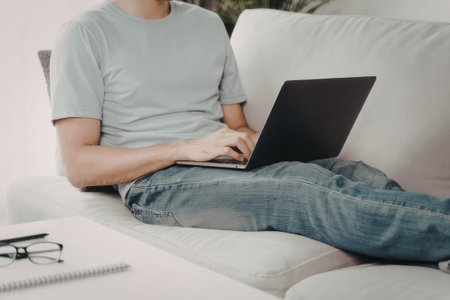 A person is comfortably seated on a white couch, using a laptop computer. They are casually dressed...