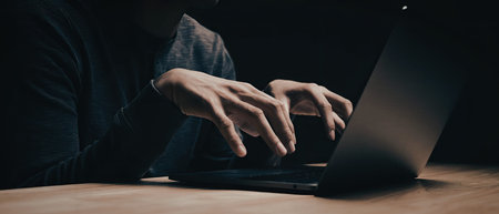 A close-up image of a person using a laptop in a dimly lit environment. The focus is on the...