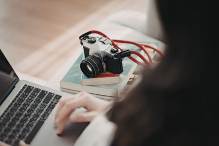 A person is seen working on a laptop with a camera and several books on the desk.