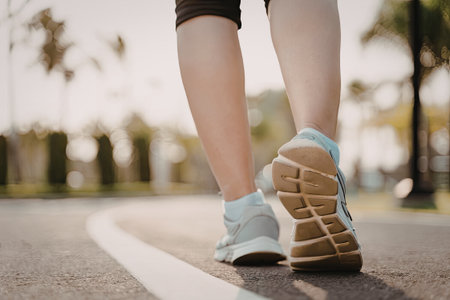 A close-up view of a person's legs and feet walking on a paved pathway in a park. The image captures the motion of walking with the person wearing white sneakers and black shorts. The background shows a blurred view of trees and greenery.の写真素材