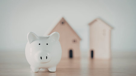 A white piggy bank sits in the foreground with two miniature wooden houses in the background.の写真素材