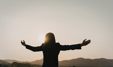 A person stands with arms outstretched against a backdrop of a setting sun, creating a silhouette. The scene is set in a natural landscape with mountains in the distance.の写真素材