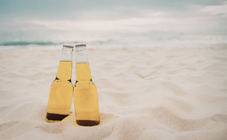 Two beer bottles standing upright in the sand on a serene beach with gentle waves and a clear sky.の写真素材