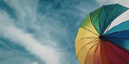 A colorful rainbow umbrella stands out against a bright blue sky with wispy clouds.の写真素材