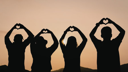 Four silhouetted individuals stand in a line against a vibrant sunset, each forming a heart shape with their hands. The warm hues of the setting sun create a serene and romantic atmosphere.の写真素材