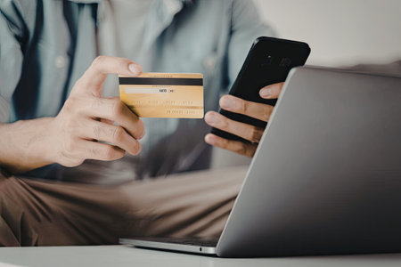 A person is holding a credit card in one hand and a smartphone in the other, with a laptop nearby....