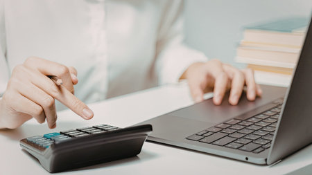 An individual is seen working on a calculator and typing on a laptop, likely performing financial...