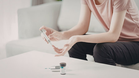 A person is sitting on a chair and testing their blood sugar level using a glucometer. The individual is holding a blood sugar testing device and there are several medical supplies on the table.の写真素材