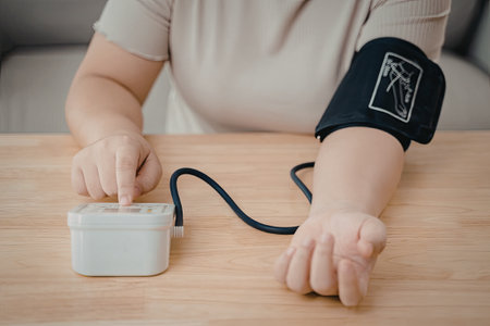 A person is using a blood pressure monitor to check their blood pressure. The individual is seated at a wooden table, focusing on the device attached to their arm.の写真素材