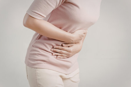 A woman wearing a pink shirt and light-colored pants is holding her stomach with both hands. She...