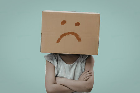 A person with a sad expression is seen wearing a cardboard box with a drawn-on sad face, arms crossed, against a light blue background.の写真素材