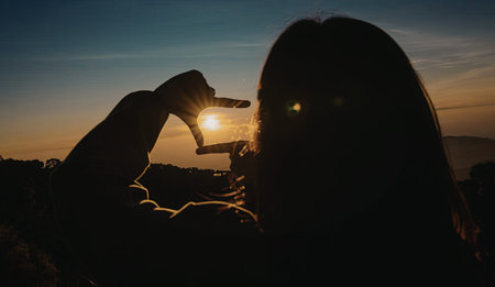 A silhouette of a person holding up their hands to frame the setting sun, with the sky displaying...