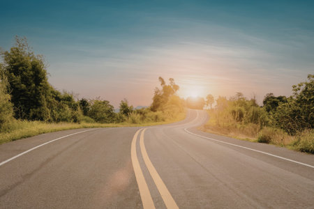 A serene image of a long, winding road stretching towards the horizon, flanked by lush greenery and bathed in the warm glow of a setting sun.の写真素材