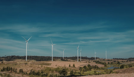 A serene landscape featuring multiple wind turbines set against a clear blue sky, surrounded by rolling hills and patches of trees.の写真素材