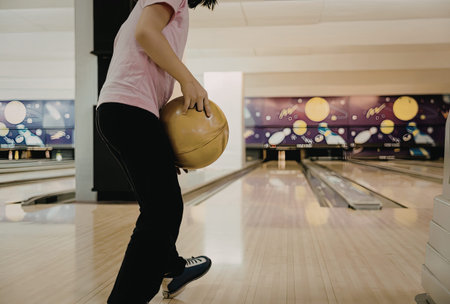 A woman in a pink shirt and black pants is holding a bowling ball, preparing to bowl in an indoor...