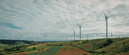A panoramic view of multiple wind turbines standing in a vast, open field. The sky is partly cloudy,...