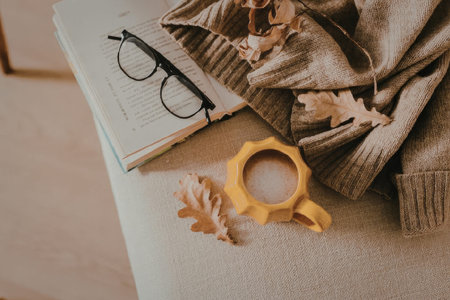 A cozy autumnal setup featuring a pair of glasses, an open book, a brown sweater, a few fallen leaves, and an orange candle holder on a beige couch.の写真素材