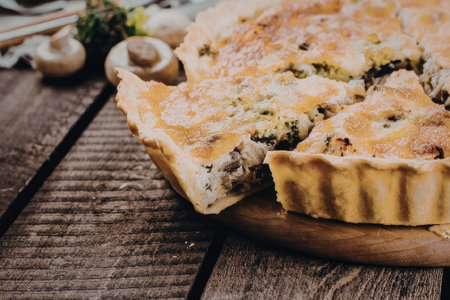 A savory quiche with a golden-brown crust and green vegetables, served on a wooden plate with mushrooms and herbs in the background.の写真素材