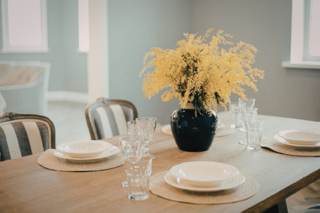 A dining room table set with plates, glasses, and a vase of yellow flowersの写真素材