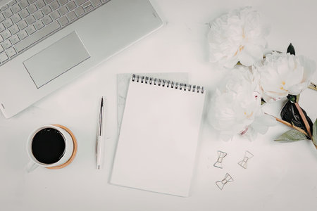A modern workspace setup with a laptop, coffee, notebook, pen, and flowers on a white desk.の写真素材
