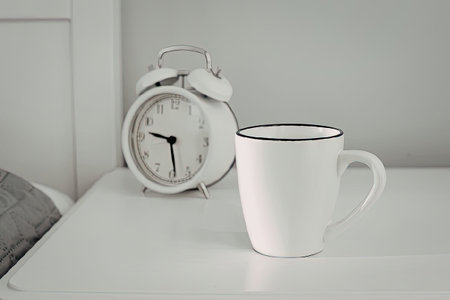 A white coffee mug on a table with an alarm clock in the backgroundの写真素材