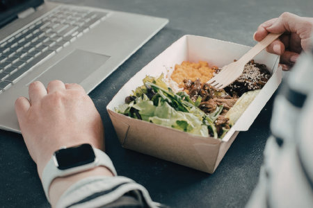 A person eating a salad from a takeout container at their desk with a laptopの写真素材