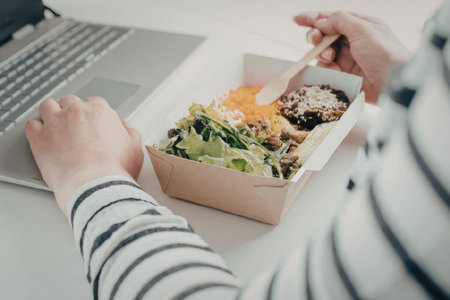 A person in a striped shirt eating a salad from a takeout container while working on a laptop.の写真素材