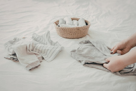 A close-up shot of a person's hands folding neutral-colored baby clothes on a white bedsheet next to a small wicker basket filled with rolled garments.の写真素材