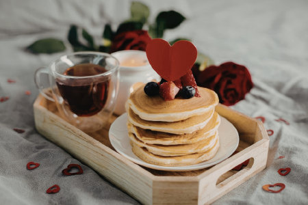 A stack of pancakes on a plate with a cup of tea and roses in the background, creating a romantic breakfast setting.の写真素材
