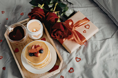 A romantic breakfast setup featuring a stack of pancakes on a wooden tray, accompanied by roses, a gift, and a candle on a bed.の写真素材