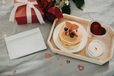 A romantic breakfast setup featuring a stack of pancakes on a wooden tray, accompanied by a heart-shaped glass of syrup, a tea light candle, a gift box, and red roses on a gray tablecloth.の写真素材