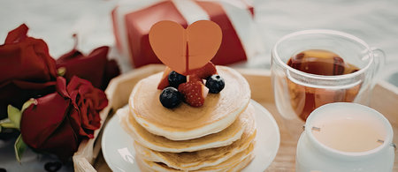A stack of pancakes with berries and a heart-shaped decoration on top, served with tea and roses.の写真素材