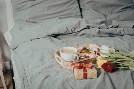 A romantic breakfast setup on a bed with a cup of coffee, a wrapped gift, and a bouquet of colorful flowers.の写真素材