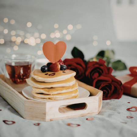 A stack of pancakes with berries and a heart on top, served on a wooden tray with a cup of tea, surrounded by red roses and heart decorations.の写真素材