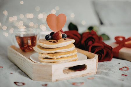 A stack of pancakes with a heart shaped decoration on top, served on a wooden tray with a cup of tea, surrounded by red roses and heart confetti.の写真素材