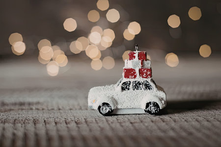 A small white toy car with presents on top, placed on a gray surface with blurred lights in the background.の写真素材