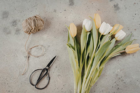 A bouquet of white and yellow tulips with green leaves, accompanied by a pair of black scissors and a ball of twine on a gray surface.の写真素材