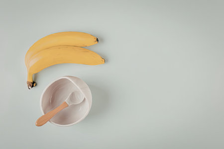 A top-down view of two bright yellow bananas placed next to a small white ceramic bowl containing a wooden spoon. The items are arranged on a clean, light blue background, suggesting a theme of healthy baby food preparation or a simple morning breakfast.の写真素材