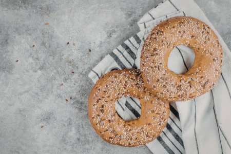 A high-angle, top-down photograph featuring two golden-brown baked bagels covered in a variety of seeds like sesame and flax. The bagels are resting on a white fabric napkin with thin black stripes, which is laid out on a textured, light grey stone or concrete surface. The scene is brightly lit, emphasizing the crunchy texture of the seeds and the soft, doughy interior of the bagels.の写真素材