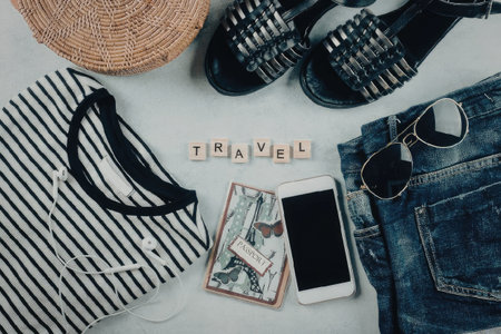 A collection of travel essentials including a bag, sandals, jeans, sunglasses, a phone, and a passport laid out on a flat surface.の写真素材
